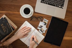 hands writing in a notebook with a laptop and coffee nearby